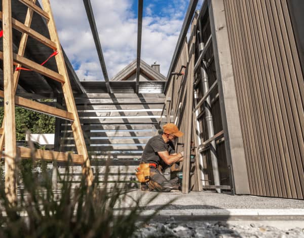 Ein Handwerker mit Werkzeugtasche arbeitet kniend an der Seitenwand eines im Bau befindlichen Carports. Sichtbar sind Metallstreben, vertikale Holzpaneele, eine Holzleiter und ein offener Dachrahmen; im Hintergrund blauer Himmel mit Wolken und Häuserdächer.