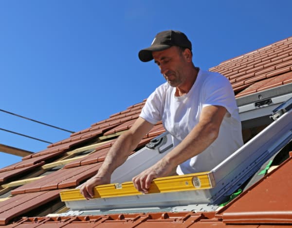 Ein Handwerker im weißen T‑Shirt steht in einer Dachöffnung und hält eine gelbe Wasserwaage an den Rahmen eines Dachfensters. Um ihn herum liegen rote Dachziegel; klarer blauer Himmel im Hintergrund. Sichtbar sind Rahmen, Befestigungselemente und die teils abgedeckte Eindeckung.