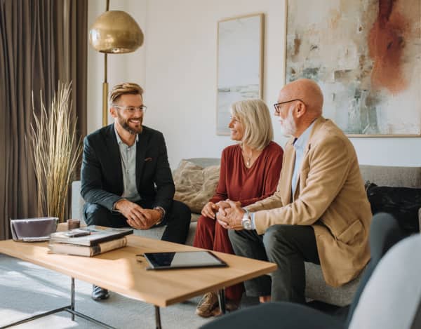 In einem modern eingerichteten Wohnzimmer sitzen drei Personen beisammen: Ein Mann im Anzug gegenüber einem Paar auf einem Sofa. Auf dem Holz-Couchtisch liegen ein Tablet, Bücher und eine Tasche; im Hintergrund stehen eine Stehlampe, Vorhänge und abstrakte Wandbilder.