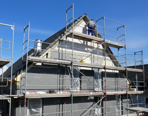 Baustellenszene bei klarem Himmel: Ein Haus mit Satteldach ist vollständig eingerüstet, während graue Dämmplatten an der Fassade angebracht werden. Ein Handwerker arbeitet am Giebel, Leitern und Baumaterial liegen auf den Gerüstebenen; Fensteröffnungen sind mit Folie abgedeckt.