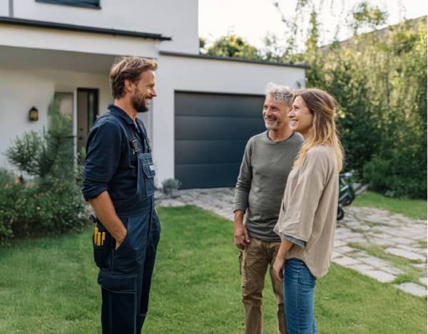 Tageslichtaufnahme vor einem modernen Einfamilienhaus mit dunklem Garagentor: Ein Handwerker in dunkler Arbeitskleidung steht links, rechts gegenüber zwei Personen in Freizeitkleidung. Im Vordergrund ist eine gepflegte Rasenfläche mit Steinweg, im Hintergrund Büsche und der Hauseingang sichtbar.