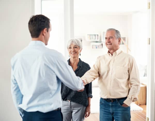 Ein älteres Paar steht in einem hellen Wohnzimmer; der Mann schüttelt einem jüngeren Mann in Hemd die Hand. Die Frau steht daneben und schaut zu. Im Hintergrund sind Regal, Sofa und Wohnaccessoires zu sehen. Helle, freundliche Innenraumatmosphäre.