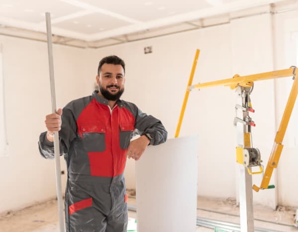 Ein Handwerker in grau-roter Arbeitskleidung steht in einem hellen, unfertigen Raum. Er hält ein Metallprofil und stützt sich auf eine Gipskartonplatte. Daneben befindet sich ein gelbes Trockenbau-Hebelgerät, im Hintergrund sind weiße Wände und eine abgehängte Decke zu sehen.
