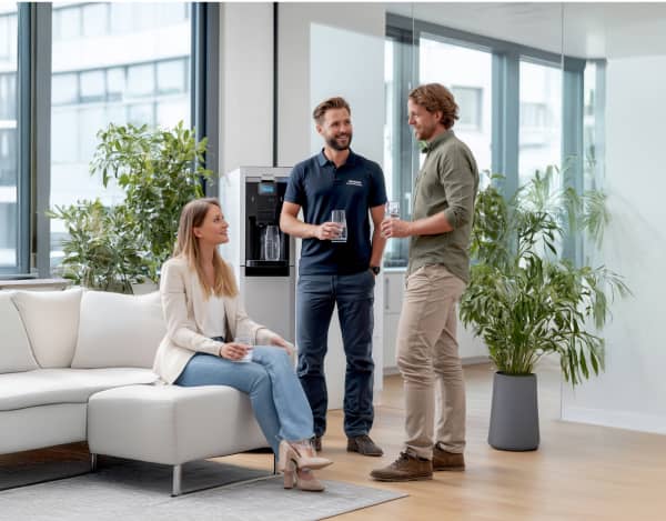 Helles Büro mit großen Fenstern, Pflanzen und heller Sofaecke. Drei Personen stehen bzw. sitzen neben einem freistehenden Wasserspender und halten Trinkgläser in der Hand. Holzfußboden und Glaswand schaffen ein freundliches, modernes Umfeld.