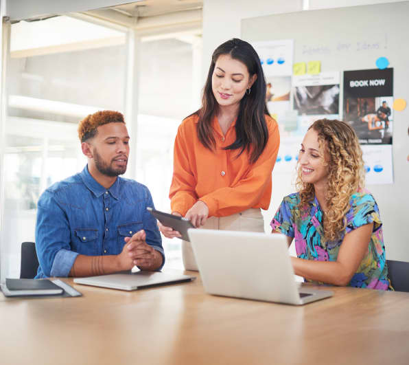 Drei Personen in einem modernen Büro betrachten gemeinsam ein Tablet; zwei sitzen mit Laptops, eine steht und präsentiert.