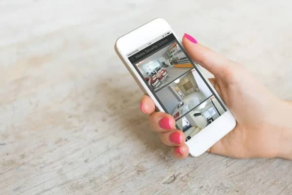 Weibliche Hand hält ein Smartphone, auf dem die Videoüberwachung verschiedener Wohnräume zu sehen ist