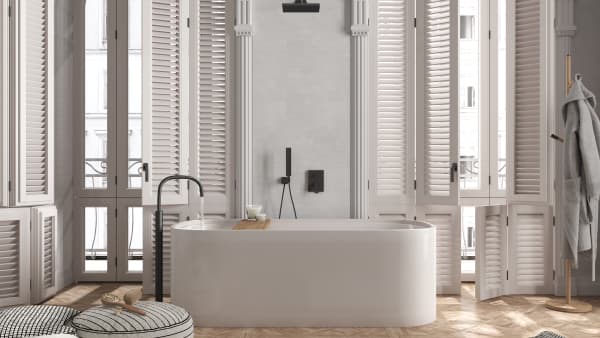 Das Bild zeigt ein stilvolles Badezimmer mit einer freistehenden Badewanne, hellen Fensterläden und modernen schwarzen Armaturen. Das Tageslicht fällt durch große Fenster, während ein Bademantel und Accessoires für eine wohnliche Atmosphäre sorgen.