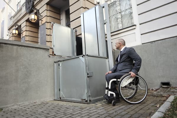 Rollstuhlfahrer vor einem Rollstuhllift vor dem Eingangsbereich eines Gebäudes.