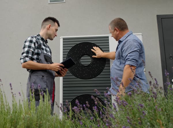 Zwei Männer betrachten eine Wärmepumpe vor einem Haus.