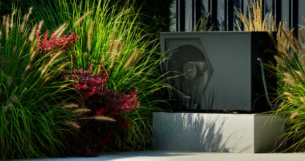 Schwarze Wärmepumpe auf Betonsockel steht vor einem Zaun in einem Garten
