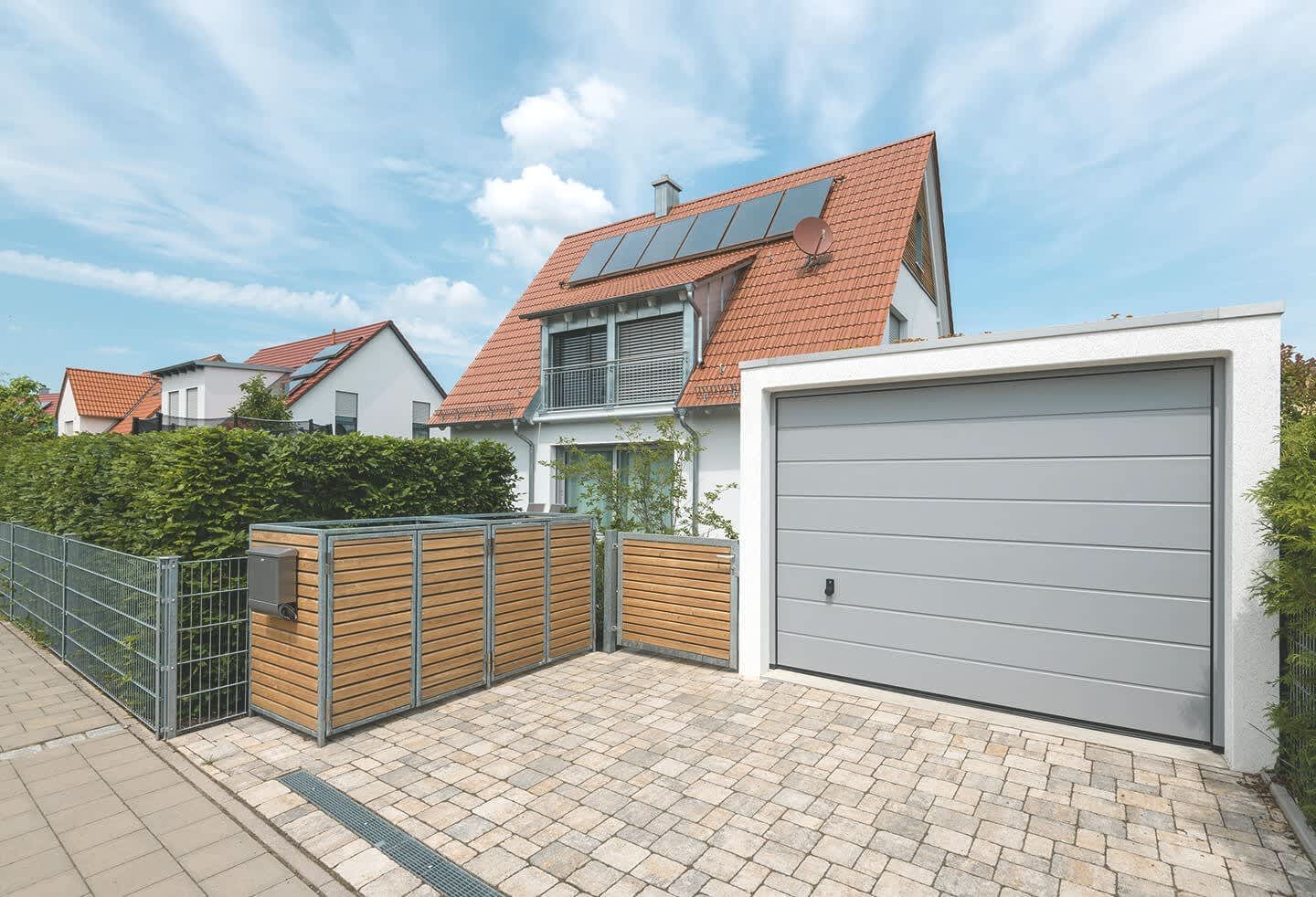 Massive, weiße Garage mit hellgrauem Tor schräg vor einem weißen Einfamilienhaus unter blauem, leicht bewölktem Himmel. Davor befindet sich eine gepflasterte Zufahrt. Hinter einer Hecke ist ein weiteres Wohnhaus zu sehen.