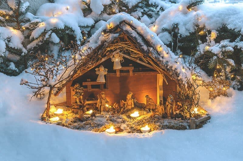 Eine beleuchtete Krippe steht im Schnee, im Hintergrund sind schneebedeckte Zeige von Nadelbäumen zu sehen