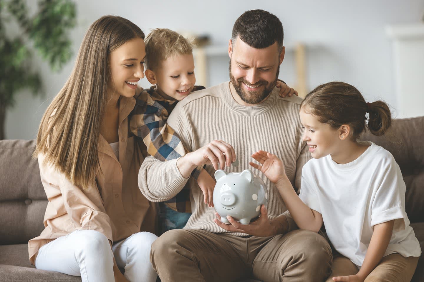 Mutter, Vater und zwei Kinder sitzen zu Hause auf dem Sofa und stecken fröhlich eine Münze ins Sparschwein.