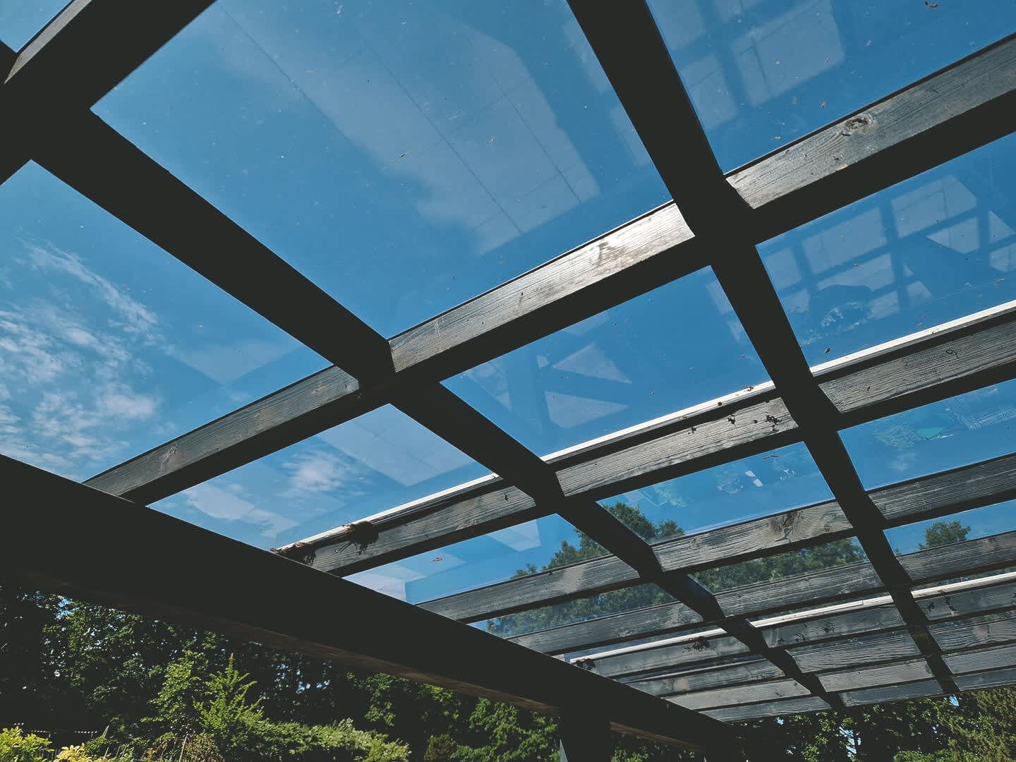 Nahaufnahme eines Terrassendachs aus Glas, das von einem stabilen Holzrahmen getragen wird. Durch die transparente Überdachung fällt Tageslicht, während der Blick auf den blauen Himmel frei ist.