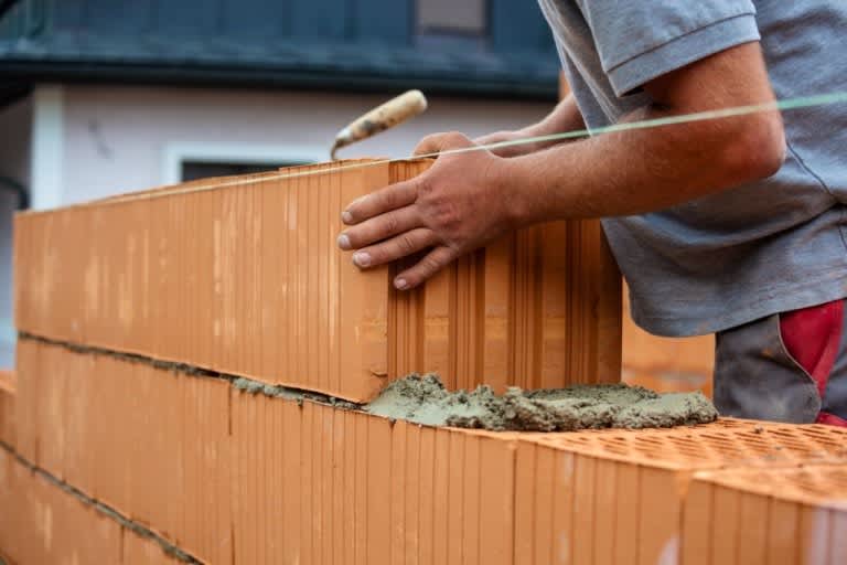 Ein Arbeiter setzt einen weiteren roten Mauerstein auf eine gemauerte Garagenwand.