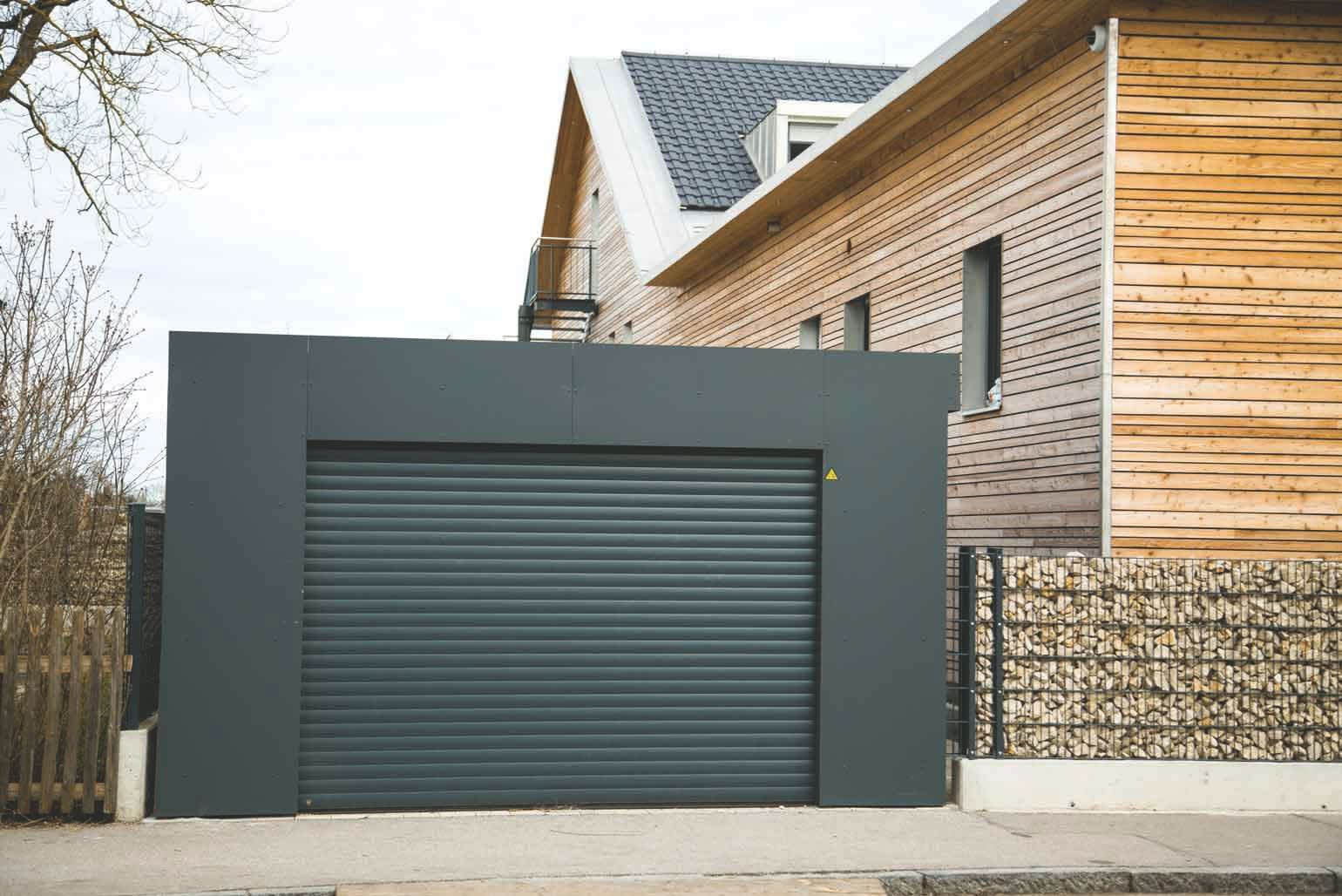 Moderne Garage aus anthrazitfarbenem Blech und Garagentor in gleicher Farbe neben einem Haus mit Holzfassade