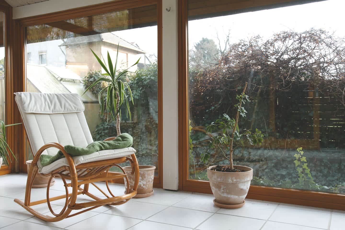 Ein Wintergarten mit großen Holzrahmenfenstern, weiß gefliestem Boden, einem Schaukelstuhl und dekorativen Zimmerpflanzen.