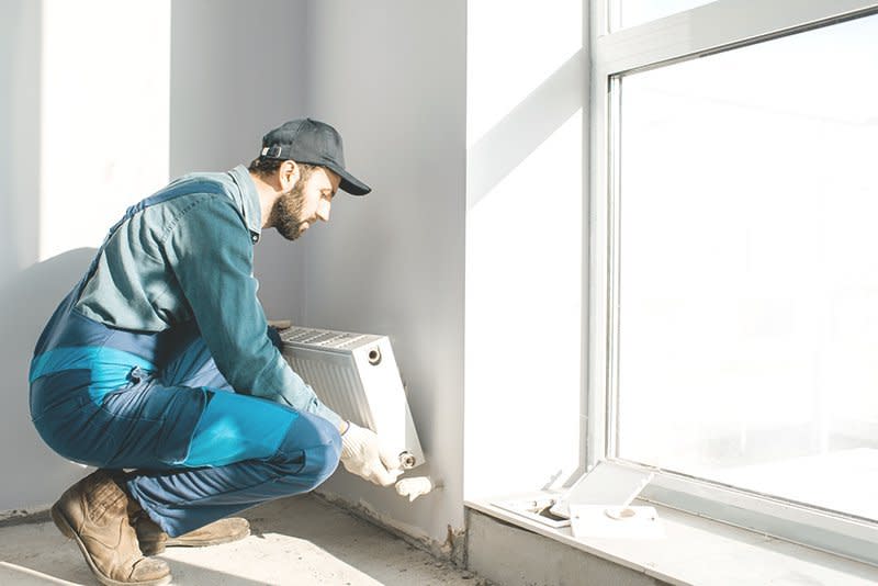 Ein Handwerker installiert einen Heizkörper neben einem großen Fenster in einem hellen Raum eines Neubaus.