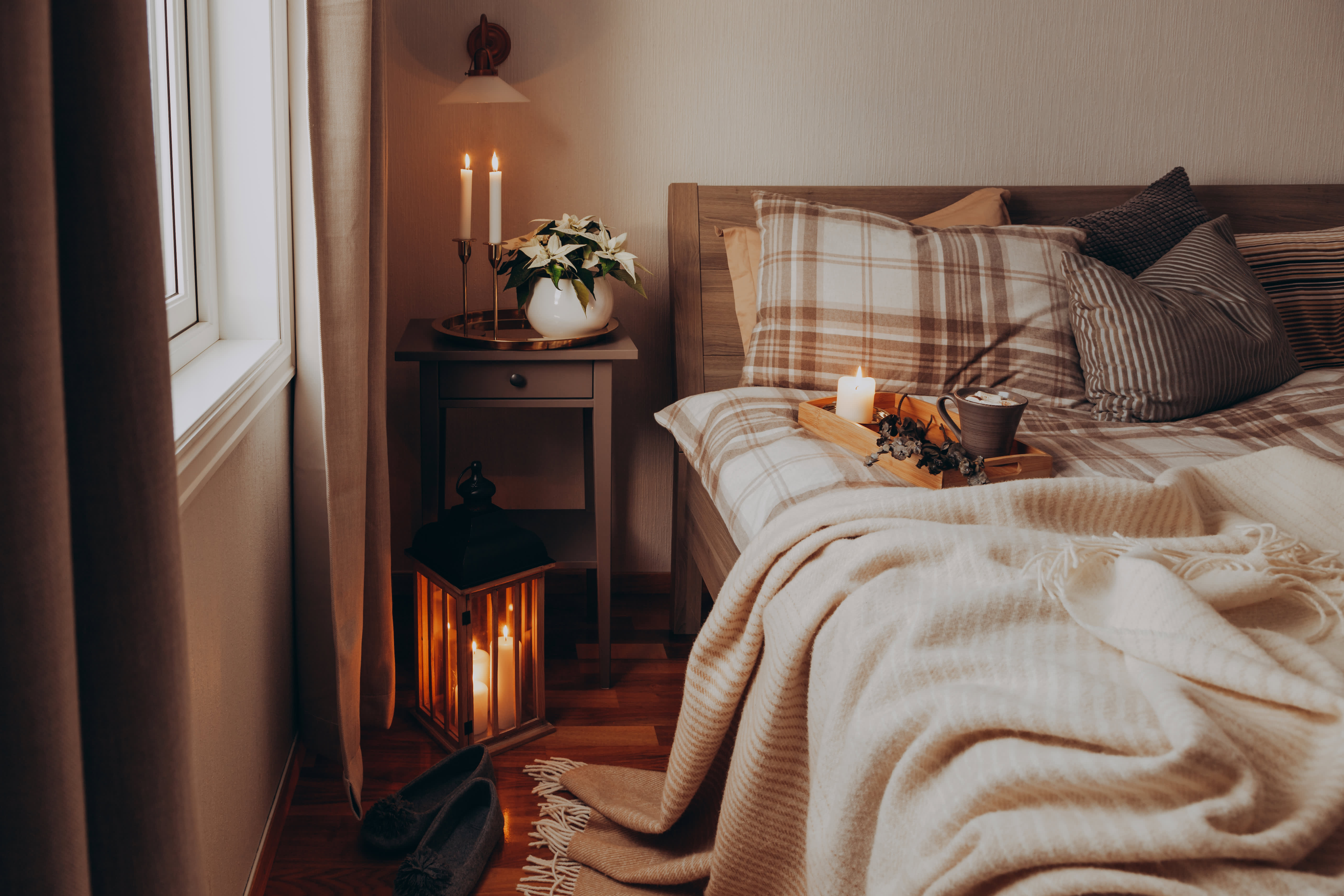 Schwach beleuchtetes Schlafzimmer, in dem ein Bett mit braun karierter Bettwäsche steht, auf dem ein Tablett mit Kerze und einer Tasse liegt.