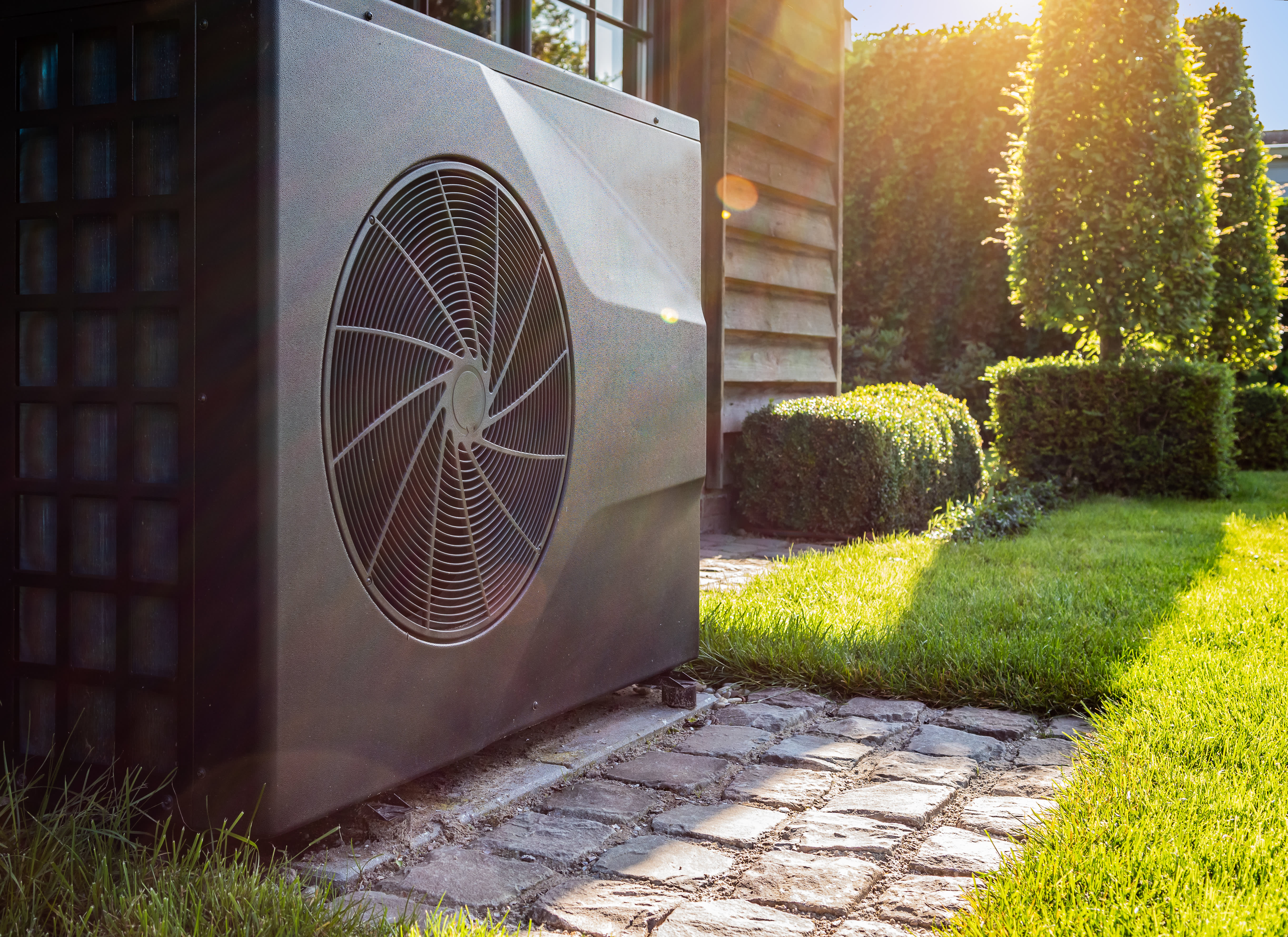 Bild einer Wärmepumpe neben einem Haus, umgeben von einem gepflegten, sonnigen Garten mit Hecken und Rasen.