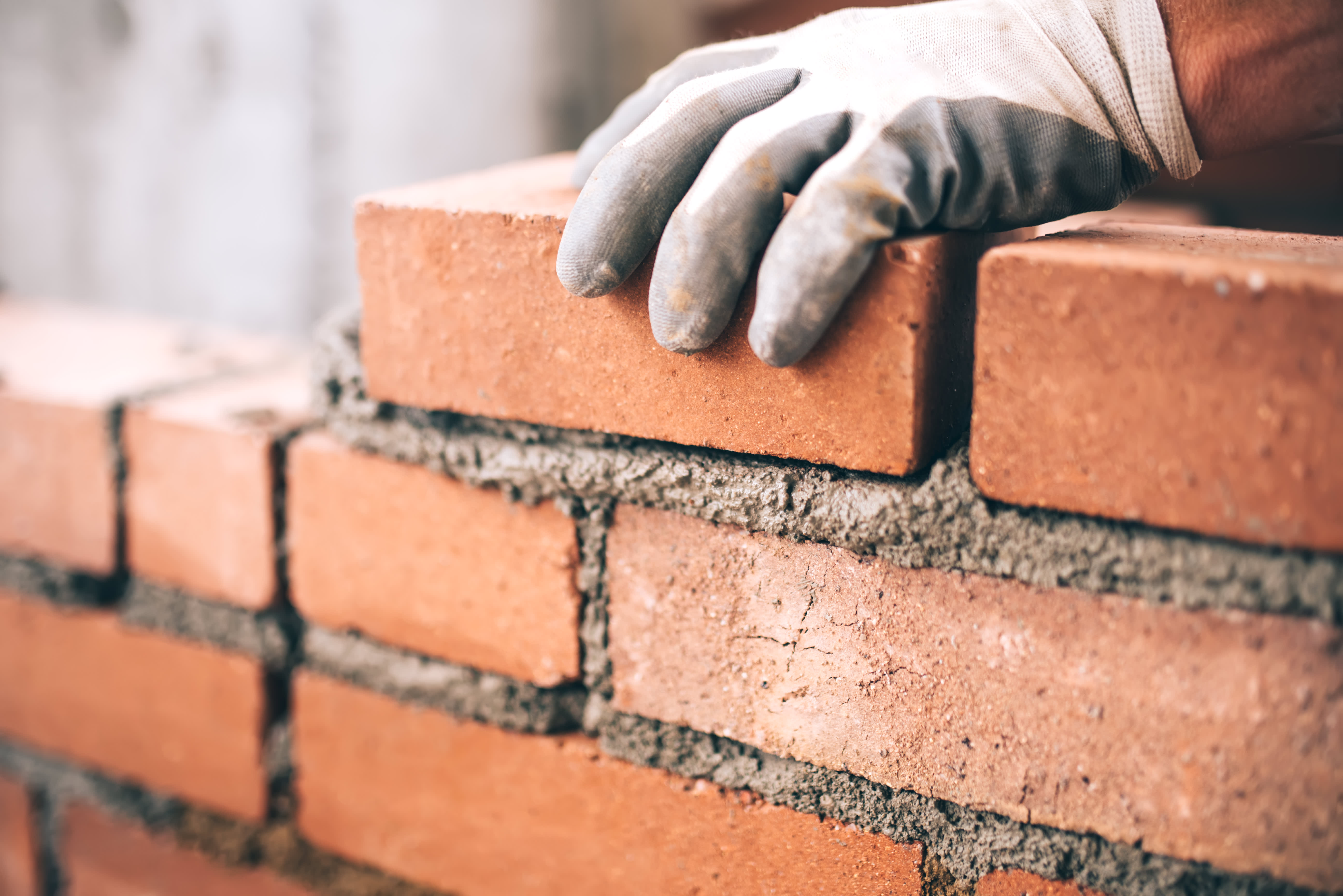 Nahaufnahme einer Mauer aus Ziegelsteinen, eine Hand mit Handschuh legt einen Ziegel