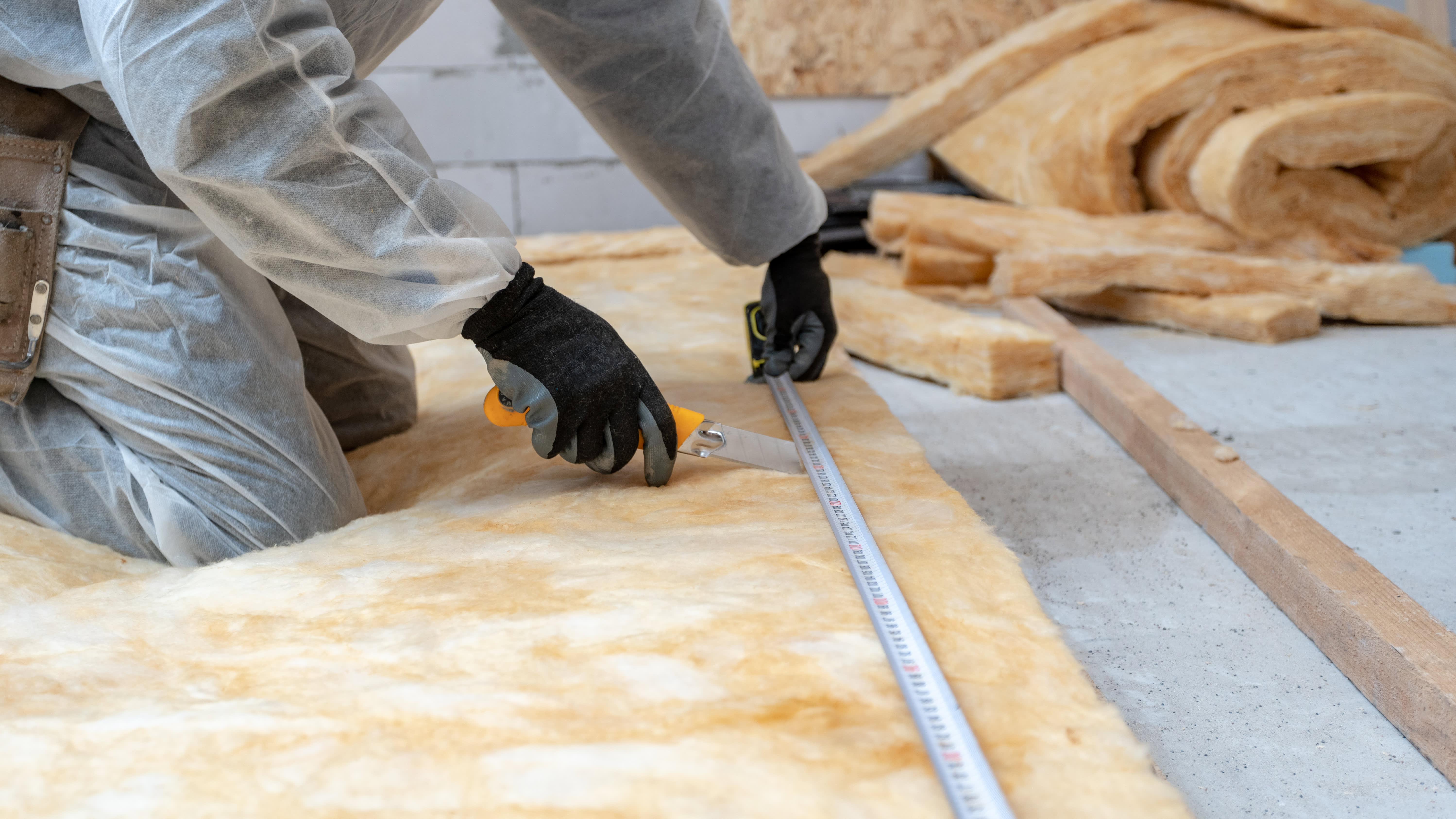 Arbeiter mit Schutzanzug kniet auf einer Matte aus Steinwolle, die er dabei zuschneidet