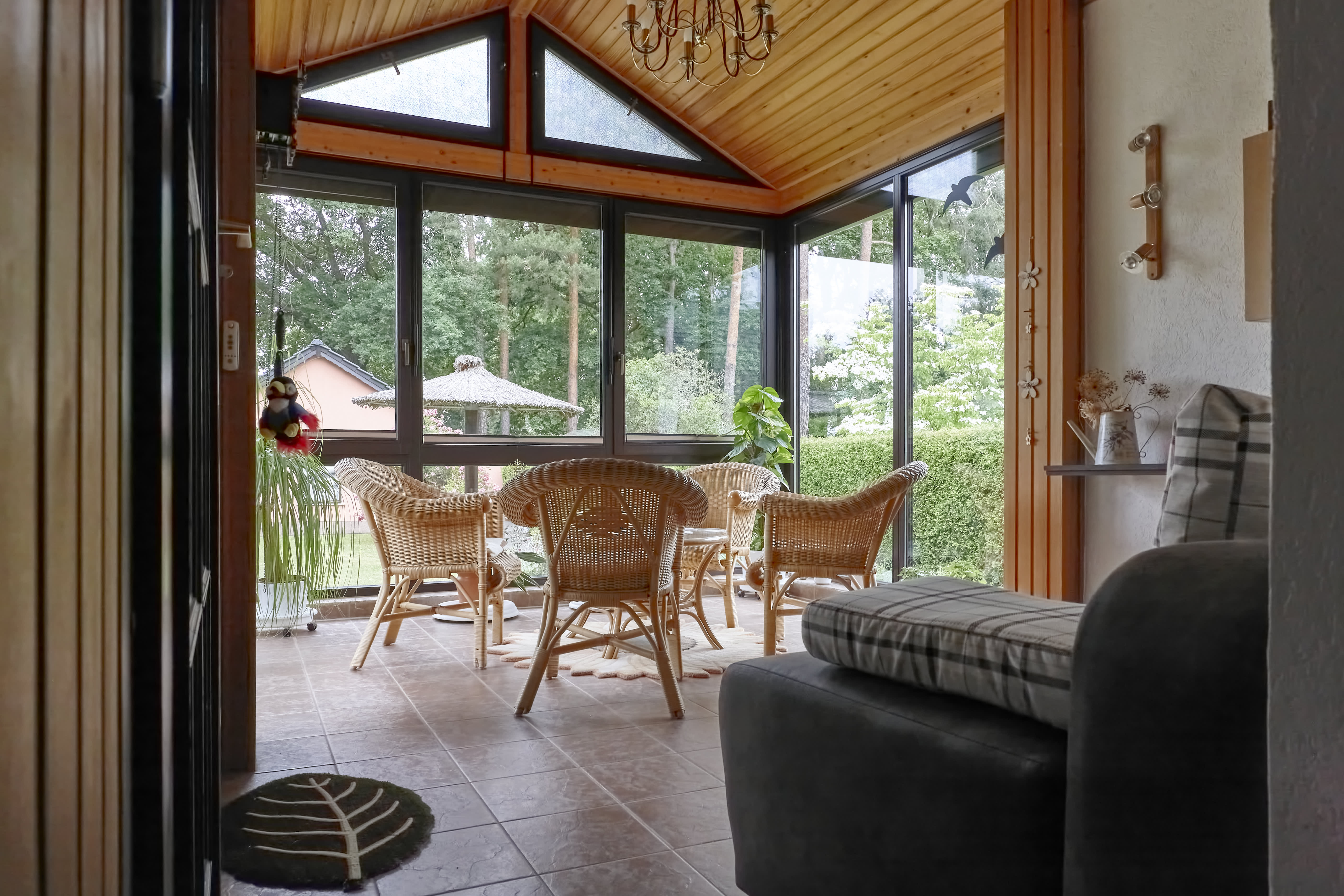 Wintergarten mit Holzdecke, großen Glasfenstern, Rattanmöbeln, Fliesenboden und Blick ins Grüne.