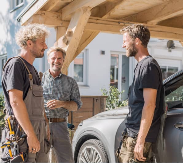 Unter einem massiven Carport aus hellem Holz stehen drei Männer und unterhalten sich. Daneben ist ein graues Auto zu sehen, im Hintergrund ein weißes Wohnhaus mit Fenstern und Gartenbereich bei sonnigem Wetter.