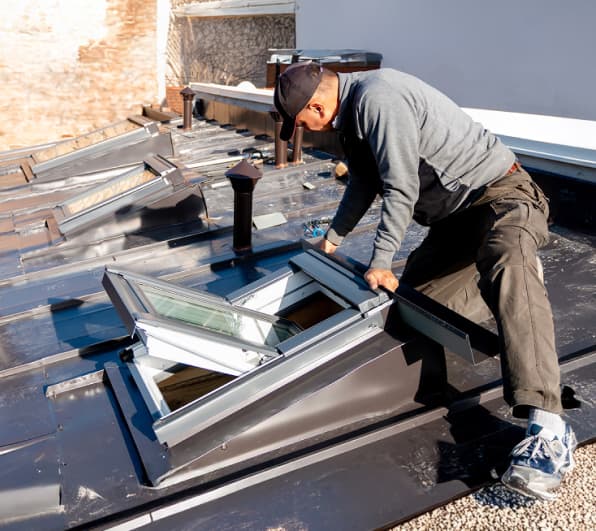 Ein Handwerker in Arbeitskleidung kniet auf einem Flachdach und richtet ein geöffnetes Dachfenster in einer Aufsatzkonstruktion aus. Um ihn herum liegen Werkzeuge; auf dem Dach sind weitere Aufbauten und Lüftungsrohre zu sehen. Helles Tageslicht, moderne Metall- und Glasflächen.