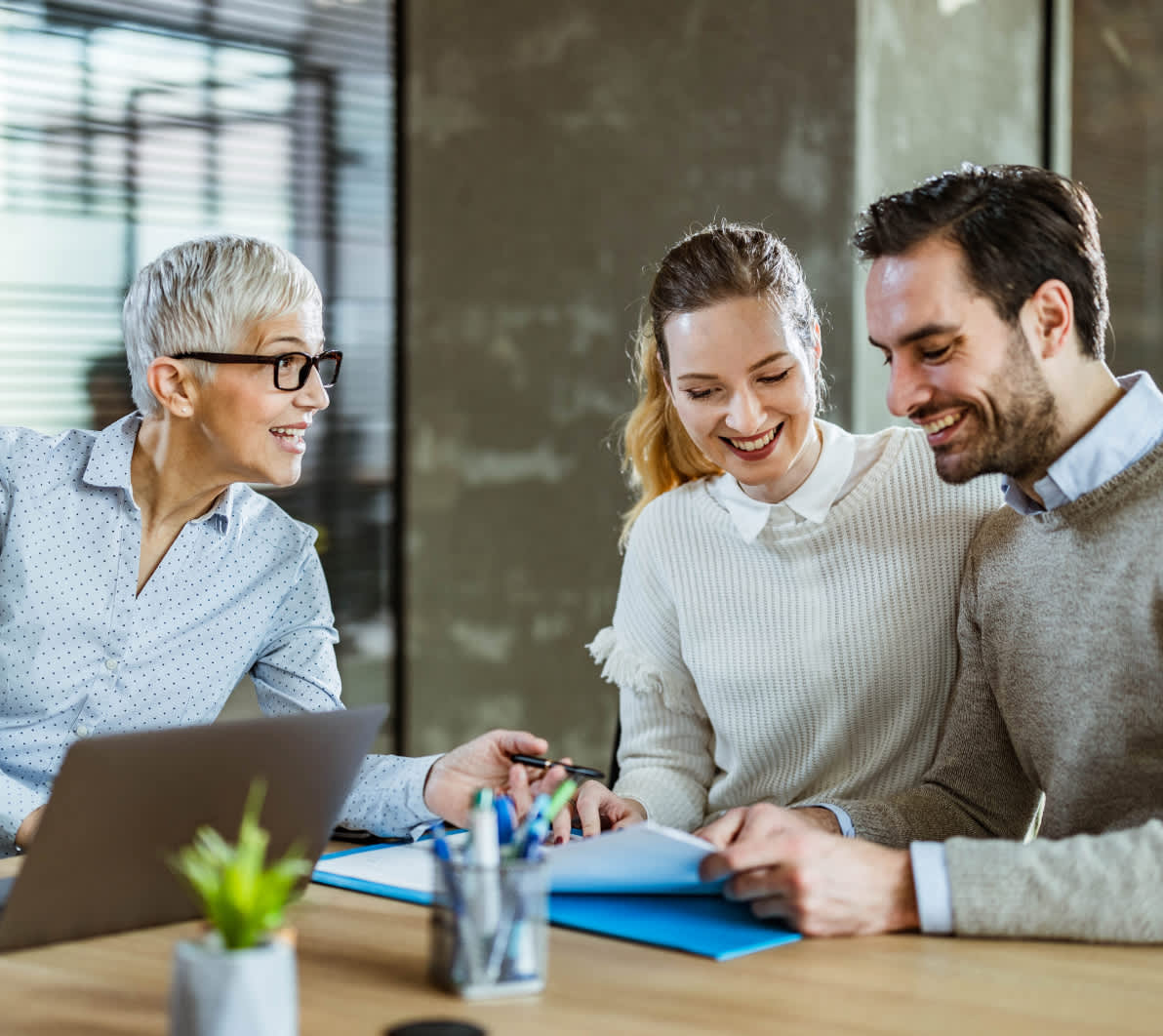 Innenaufnahme eines modernen Büros: Eine ältere Person mit Kurzhaarfrisur erklärt etwas und zeigt auf Dokumente, zwei jüngere Personen sitzen daneben und hören zu. Auf dem Holztisch liegen Ordner, Stifte und ein Notizbuch, daneben steht ein geöffneter Laptop und eine kleine Topfpflanze. Licht fällt durch große Fenster mit Lamellen in den Raum.