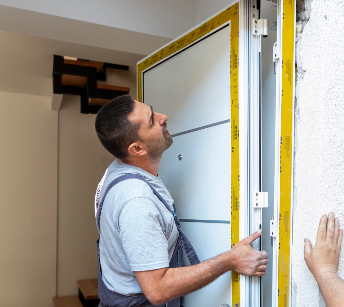 Ein Handwerker in Arbeitskleidung setzt eine weiße Haustür mit horizontalen Zierlinien in einen gelben, markierten Türrahmen ein. Nahaufnahme der Ausrichtung; neben dem Rahmen ist eine Hand sichtbar, im Hintergrund eine Treppe und Innenraum. Gesichtszüge sind unkenntlich gemacht.