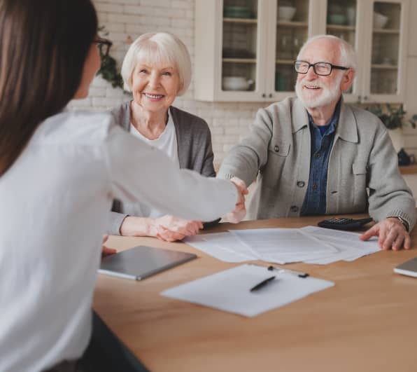 Ein älteres Paar sitzt an einem Holztisch in einem hellen Raum; der Mann reicht einer Frau mit Brille die Hand. Auf dem Tisch liegen Dokumente, Stift, Tablet und ein Taschenrechner. Neben dem Mann sitzt eine ältere Frau und schaut zu; im Hintergrund sind Schränke und Dekoration zu sehen.