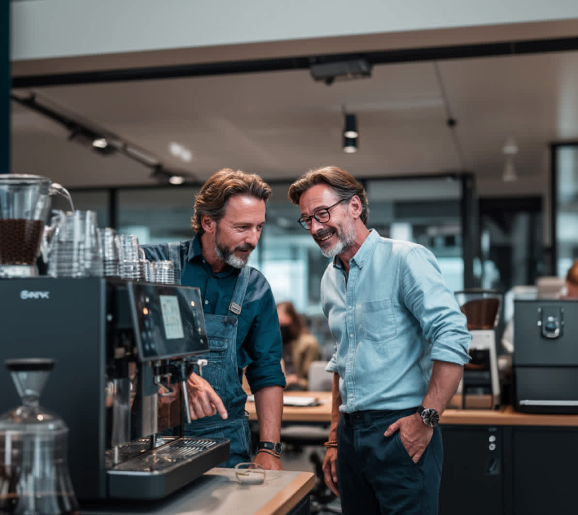 Moderner Kaffeevollautomat auf einer Theke in einem offenen Büro. Zwei Personen stehen daneben und unterhalten sich, während Tassen und Zubehör bereitstehen. Helles, professionelles Arbeitsumfeld mit Fokus auf die Kaffeemaschine und die Zubereitung.