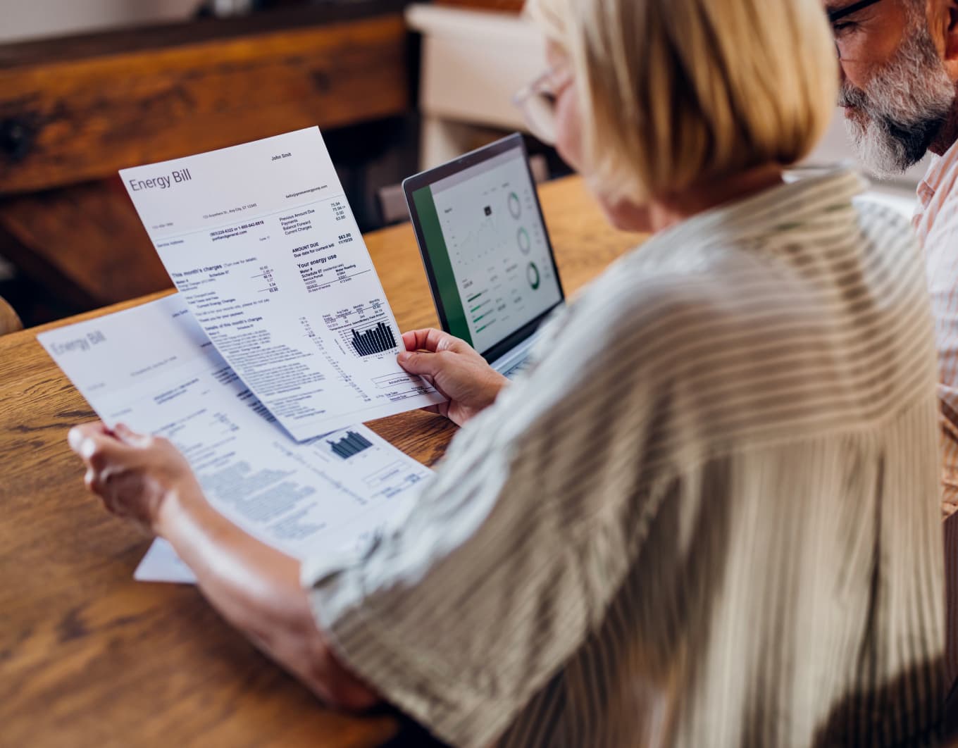 Nahaufnahme von zwei Personen an einem Holztisch: Eine Person hält mehrere Blätter mit der Aufschrift „Energy Bill“ und Diagrammen in der Hand, daneben liegt weitere Post. Im Hintergrund steht ein aufgeklappter Laptop mit geöffneten Auswertungs-Grafiken auf dem Bildschirm.