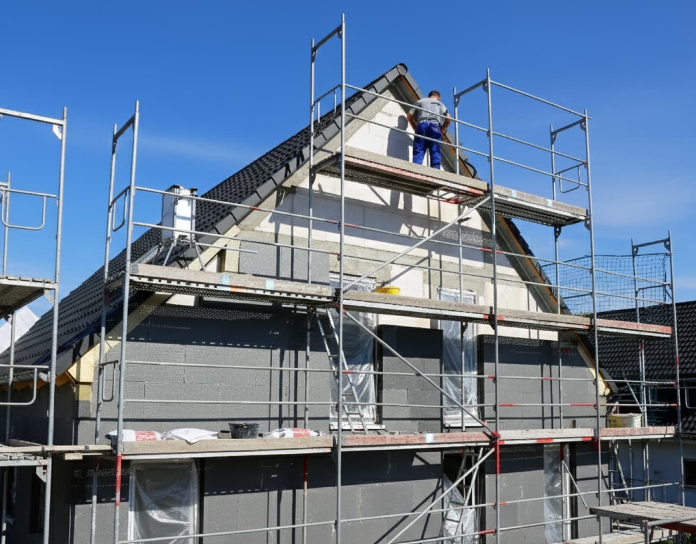Baustellenszene bei klarem Himmel: Ein Haus mit Satteldach ist vollständig eingerüstet, während graue Dämmplatten an der Fassade angebracht werden. Ein Handwerker arbeitet am Giebel, Leitern und Baumaterial liegen auf den Gerüstebenen; Fensteröffnungen sind mit Folie abgedeckt.