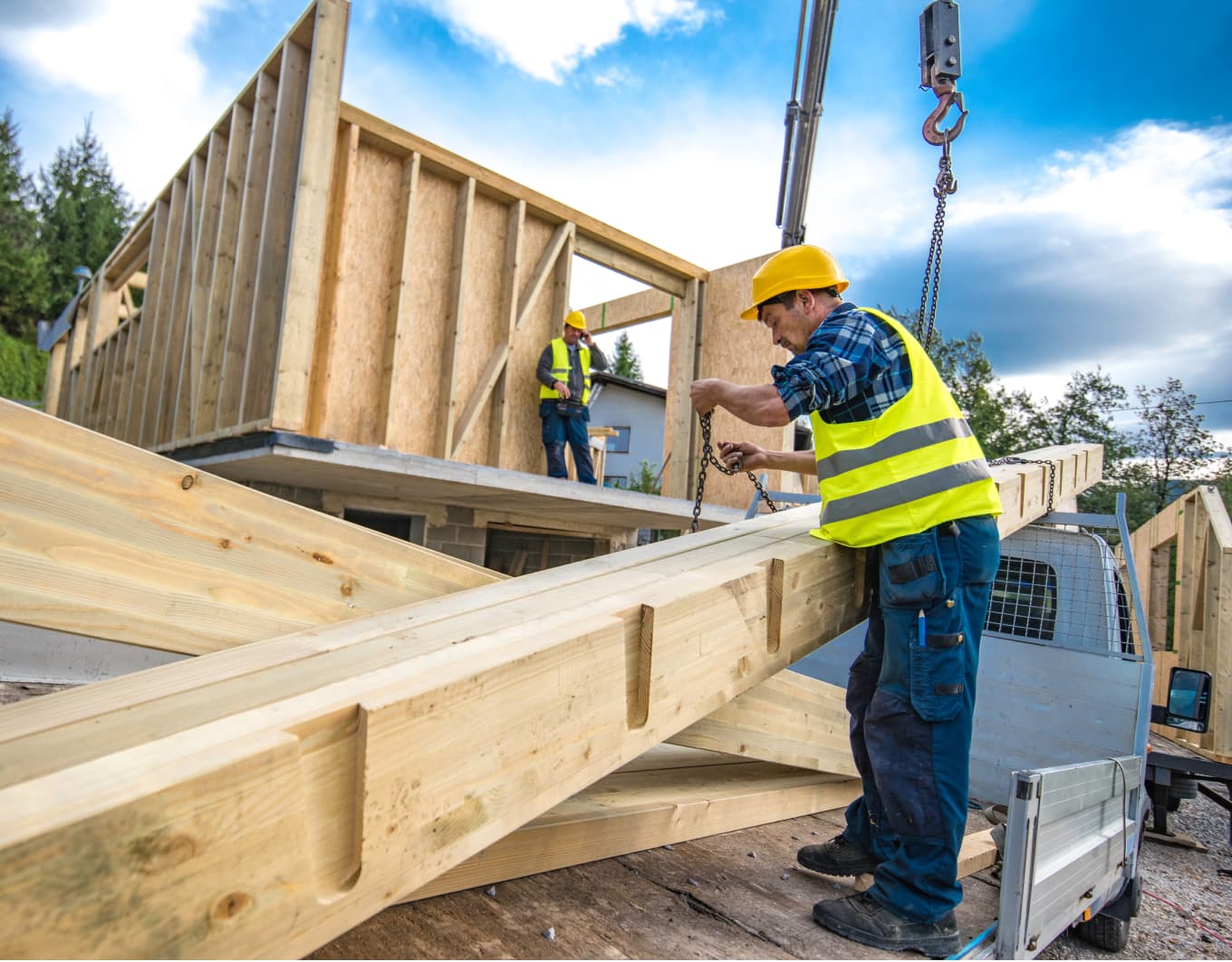 Szene auf einer Baustelle bei Tageslicht: Ein Arbeiter mit gelbem Schutzhelm und Warnweste steht auf einer Ladefläche und hängt Ketten an einen langen Holzbalken, darüber ein Haken am Kran. Im Hintergrund sind weitere Holzrahmenwände, ein zweiter Arbeiter mit Helm und Weste sowie Bäume und Himmel zu sehen.