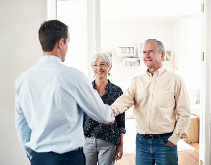 Ein älteres Paar steht in einem hellen Wohnzimmer; der Mann schüttelt einem jüngeren Mann in Hemd die Hand. Die Frau steht daneben und schaut zu. Im Hintergrund sind Regal, Sofa und Wohnaccessoires zu sehen. Helle, freundliche Innenraumatmosphäre.