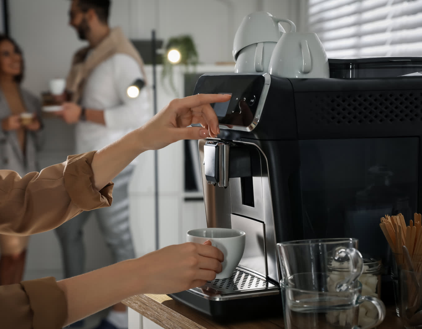 Nahaufnahme eines schwarzen Kaffeevollautomaten auf einer Theke. Eine Hand hält eine weiße Tasse unter den Auslauf und bedient das Display. Auf dem Gerät stehen Tassen, daneben stehen ein Wasserkrug und Rührstäbchen; im Hintergrund unterhalten sich zwei Personen mit Kaffeetassen.