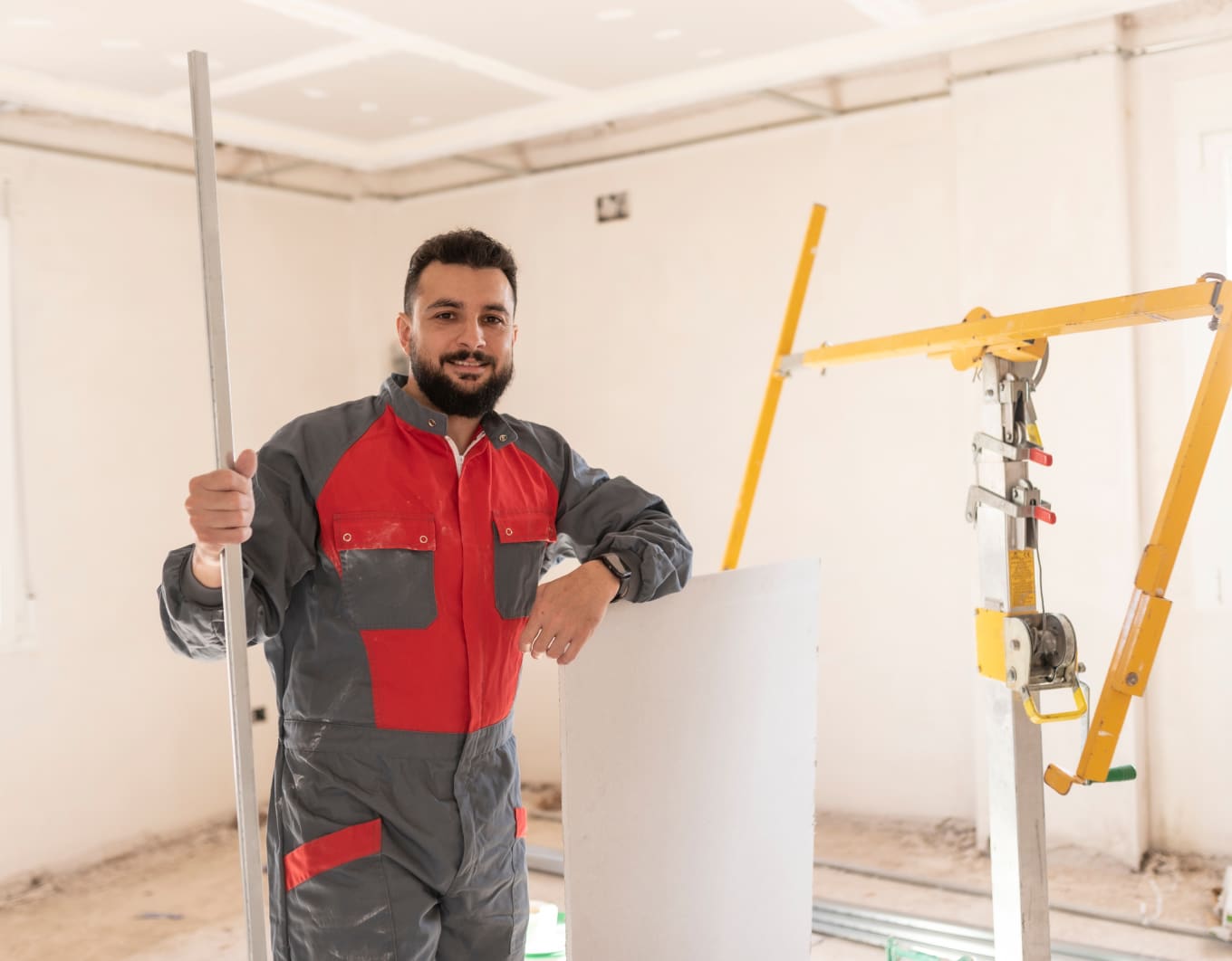 Ein Handwerker in grau-roter Arbeitskleidung steht in einem hellen, unfertigen Raum. Er hält ein Metallprofil und stützt sich auf eine Gipskartonplatte. Daneben befindet sich ein gelbes Trockenbau-Hebelgerät, im Hintergrund sind weiße Wände und eine abgehängte Decke zu sehen.