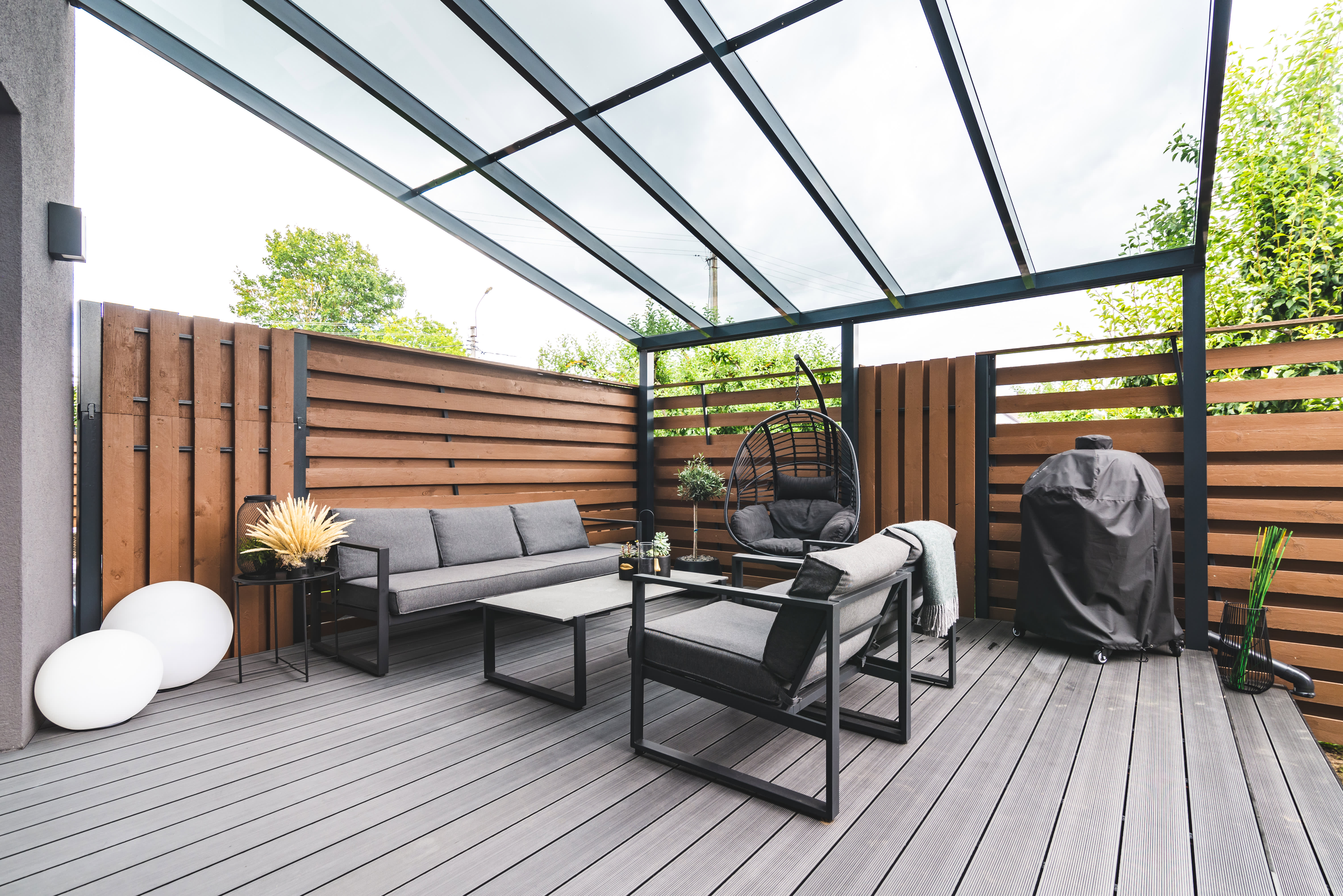 Eine überdachte Terrasse mit  transparentem Glasdach und gemütlicher Lounge-Ecke. Die Terrasse ist mit einem braunen Sichtschutzzaun vom Nachbargrundstück abgetrennt.