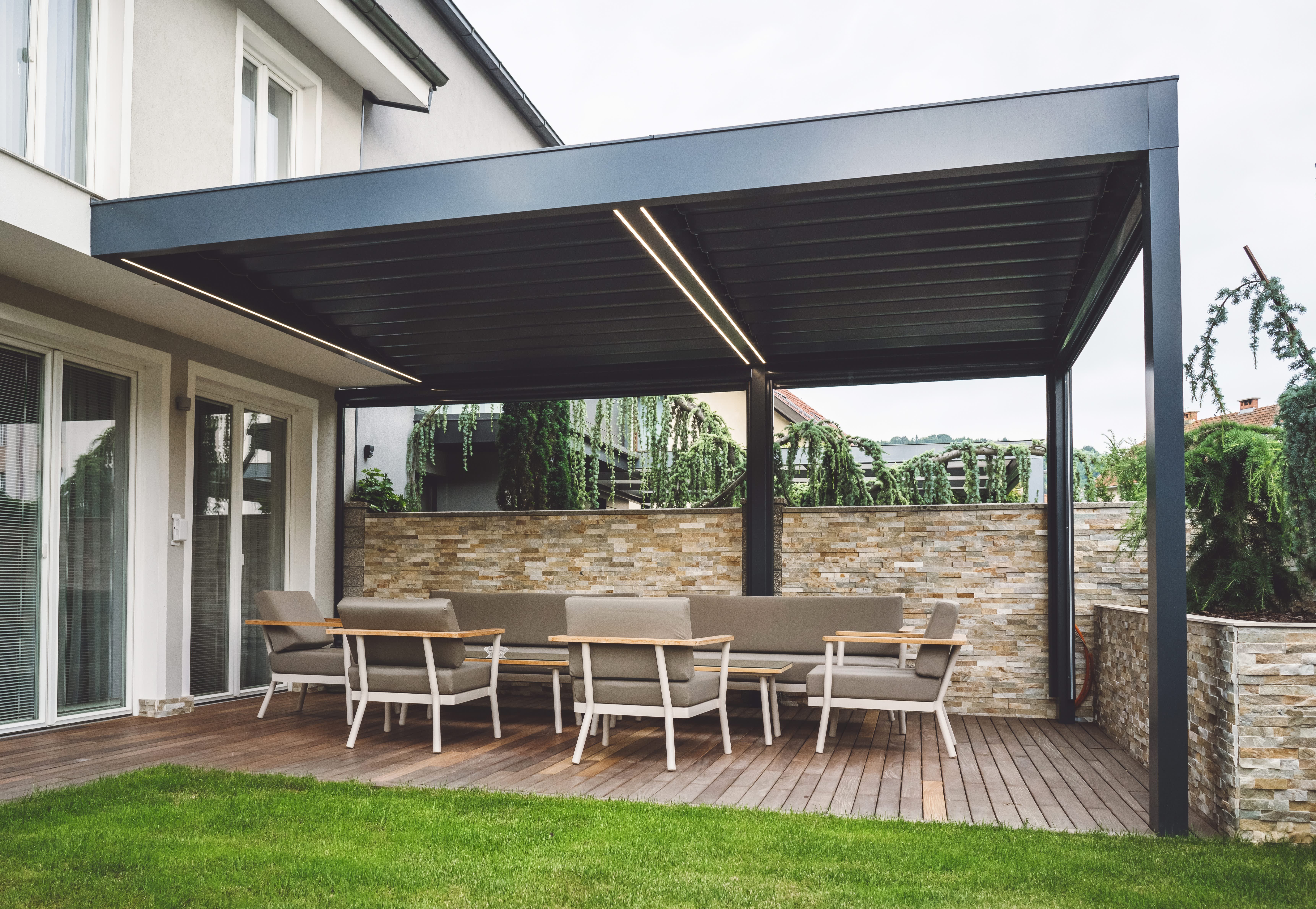 Ans Wohnhaus angebaute schwarze Terrassenüberdachung mit Beleuchtung ist über gemütlichen Loungemöbeln platziert.