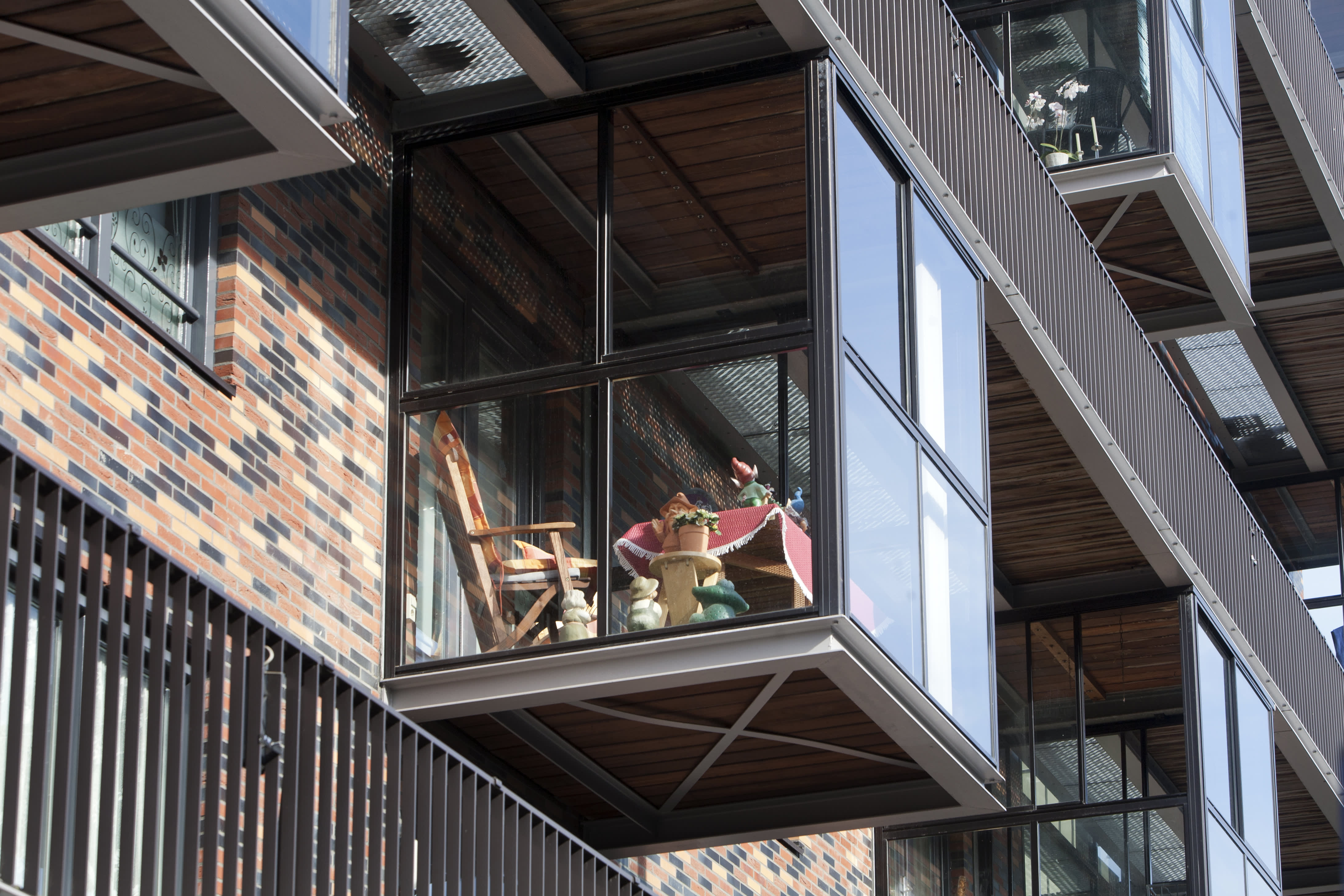 Ein moderner verglaster Balkon an einem Backsteingebäude, ausgestattet mit einem Holzstuhl, einem Tisch mit rotem Tischläufer, Topfpflanzen und kleinen Dekorationsfiguren.