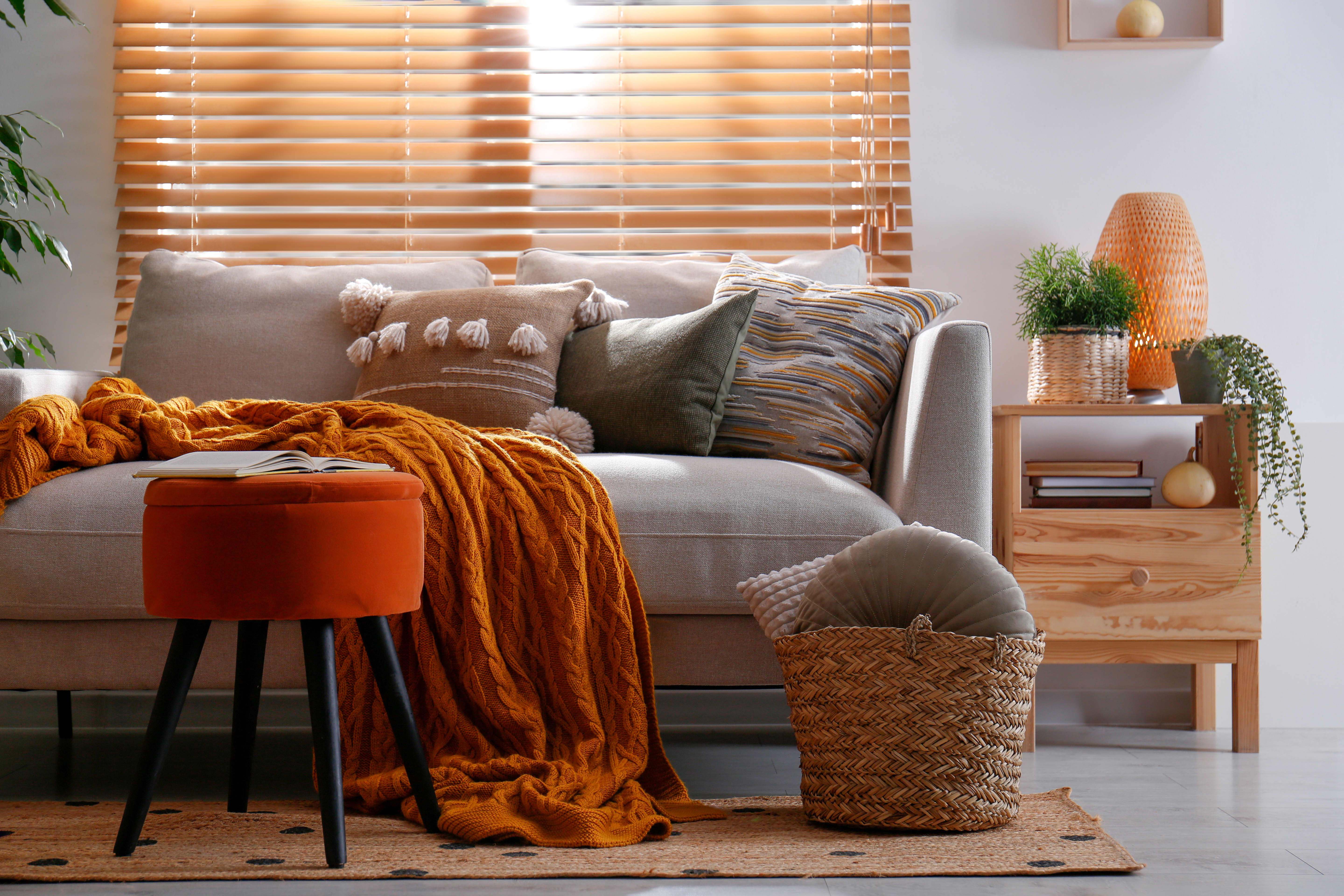 Wohnzimmer mit grauem Sofa, Beistelltisch aus hellem Holz, orangefarbenem Hocker, orangener Decke und Kissen in Grün. und Brauntönen