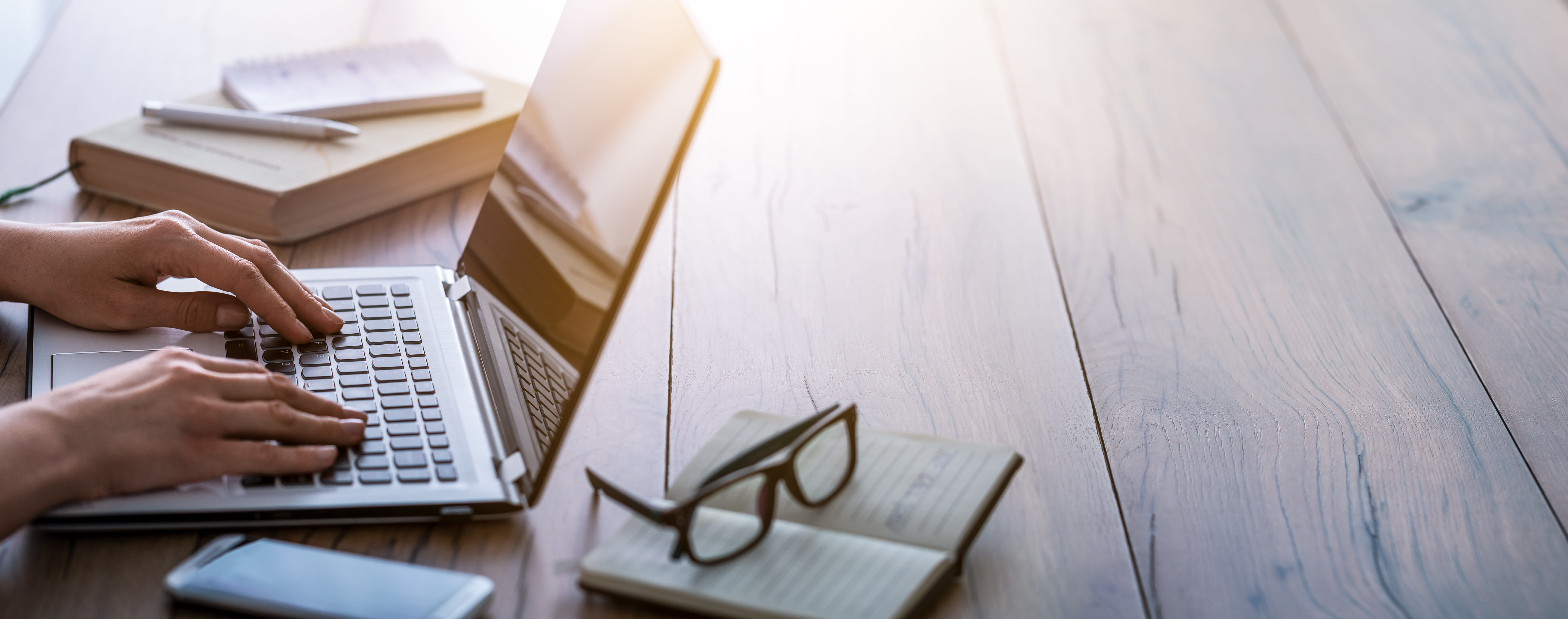 Person schreibt etwas auf dem Laptop, daneben liegen  Notizbücher und Brille