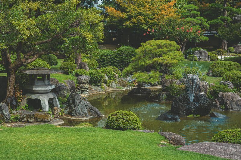 Japanischer Garten mit gepflegtem Rasen, verschiedenen Bäumen und einem Teich mit Wasserfontänen