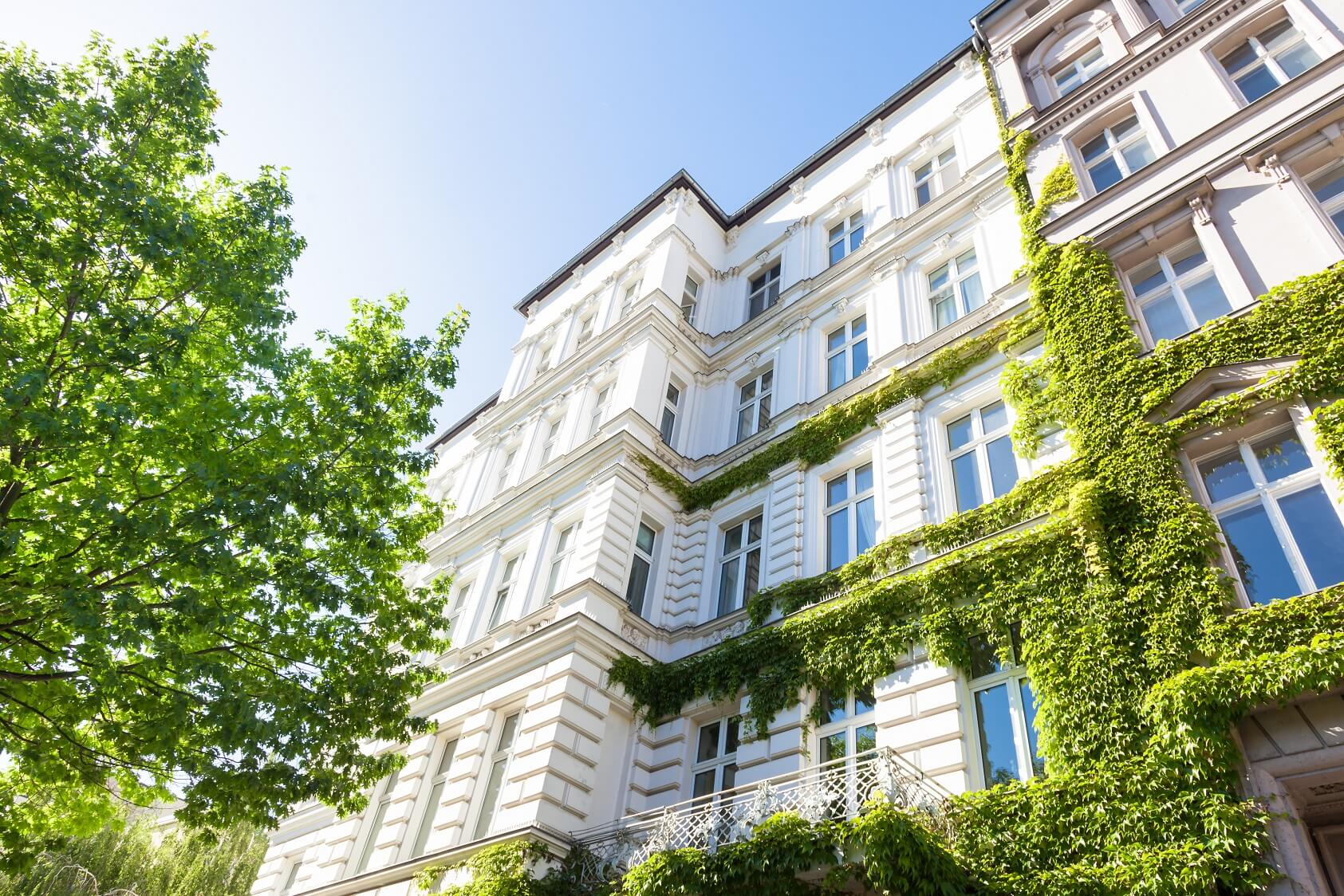 Blick von der Straße nach oben auf die weiße, teilweise mit Grünpflanzen bewachsene Fassade eines Mehrfamilienhauses